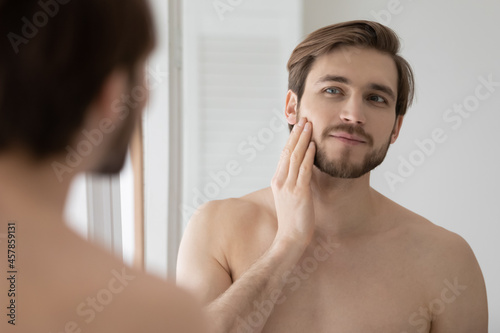 Young shirtless man looking in mirror touch his face after shave applied aftershave cream enjoy smooth, flawless, soft skin. Grooming, morning routine, ad of skincare cosmetic products for men concept photo