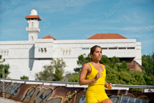 Caucasian woman in yellow sportswear running across urban bridge with mosque on the background. Horizontal panoramic view of fitness woman training outdoors.