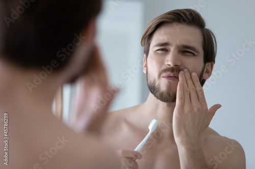 Handsome young man look in mirror touch cheek with hand, feels pain while brushing teeth due cavity or caries. Toothache, dental check up, oral care, need toothpaste for sensitive tooth enamel concept photo