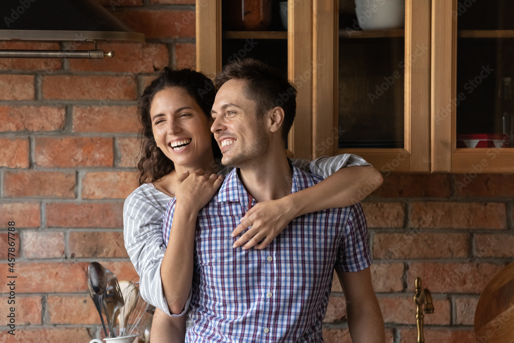Happy beautiful latin woman cuddling from back sincere smiling millennial husband, enjoying carefree weekend time together in wooden kitchen, joyful loving young family couple daydreaming indoors.