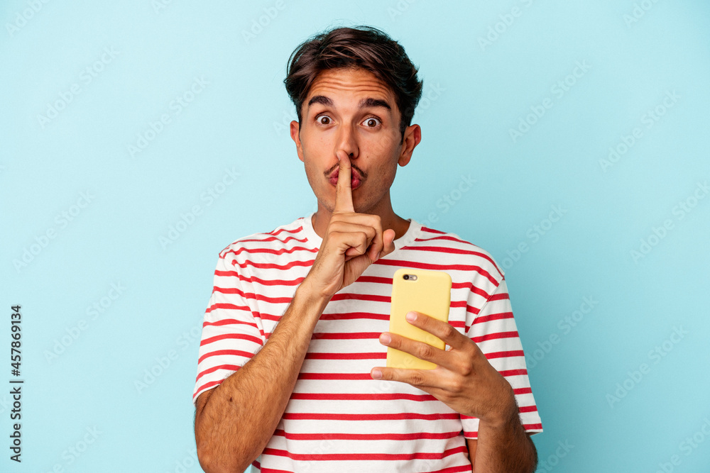 Young mixed race man holding mobile phone isolated on blue background keeping a secret or asking for silence. Young mixed race man holding mobile phone isolated on blue background keeping a secret or asking for silence.