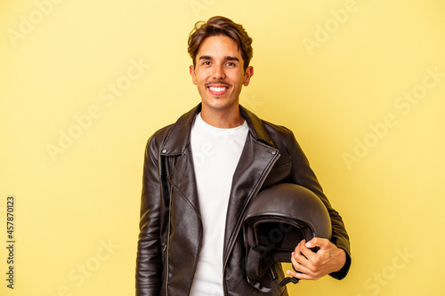 Young mixed race man holding helmet isolated on yellow background happy, smiling and cheerful.