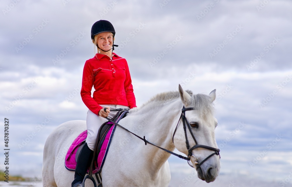 Portrait of a beautiful girl, young woman rider, equestrian on white horse in helmet and polo shirt, riding outdoors. Horseback riding sport. Portrait of a beautiful girl, young woman rider, equestrian on white horse in helmet and polo shirt, riding outdoors. Horseback riding sport.