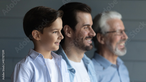 Head shot profile of three generations of men standing in row on grey studio wall background, looking in distance, little boy grandson with senior grandfather and father dreaming of good future