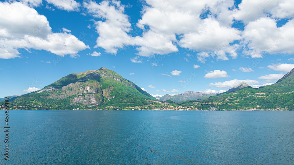 Landscape of the coastline of the famous Lake Como, Italy, with turquoise waters, green hills and Italian alps. In the distance the village of Menaggio is visible. Blue sky and white clouds. Landscape of the coastline of the famous Lake Como, Italy, with turquoise waters, green hills and Italian alps. In the distance the village of Menaggio is visible. Blue sky and white clouds.