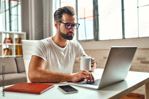 Businessman in glasses with a mug of coffee works at a laptop. Freelance, work from home.