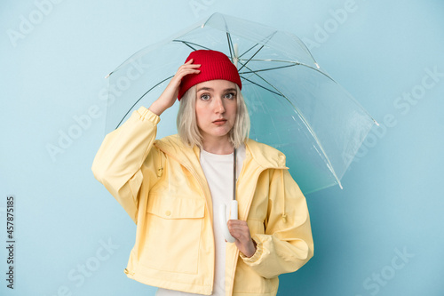 Young caucasian woman holding umbrella isolated on blue background being shocked, she has remembered important meeting.