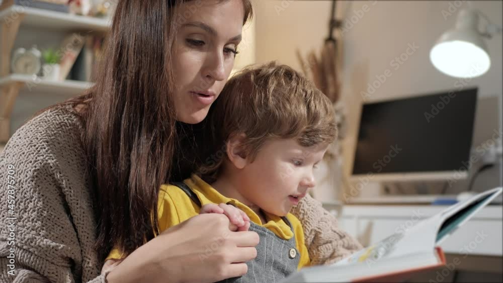 Mom reads book to baby. Mother and child are sitting on bed in evening in bedroom, mom holds boy hand and reads him book. Close-up and slow-motion Mom reads book to baby. Mother and child are sitting on bed in evening in bedroom, mom holds boy hand and reads him book. Close-up and slow-motion