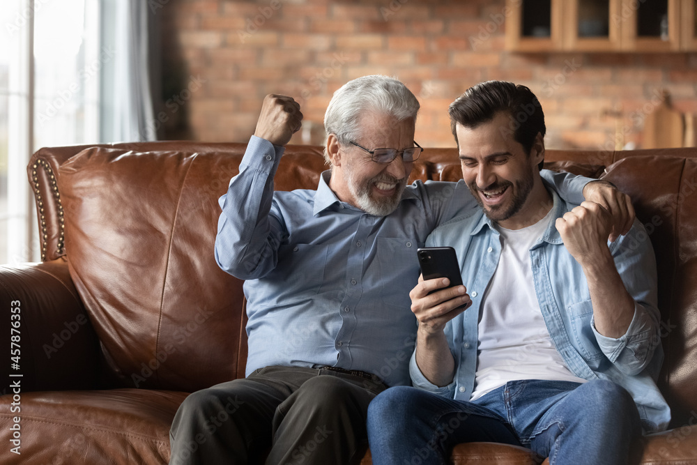 Excited two generations of men celebrating success, using smartphone, senior dad with son looking at device screen, reading good unexpected news in message, showing yes gesture, online lottery win Excited two generations of men celebrating success, using smartphone, senior dad with son looking at device screen, reading good unexpected news in message, showing yes gesture, online lottery win