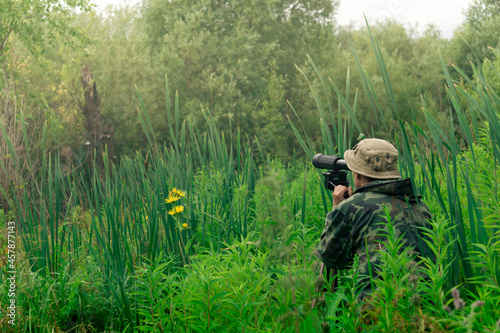 male explorer makes observations in the wild with a spotting scope standing among the tall grass