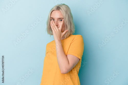 Young caucasian woman isolated on blue background blink at the camera through fingers, embarrassed covering face.