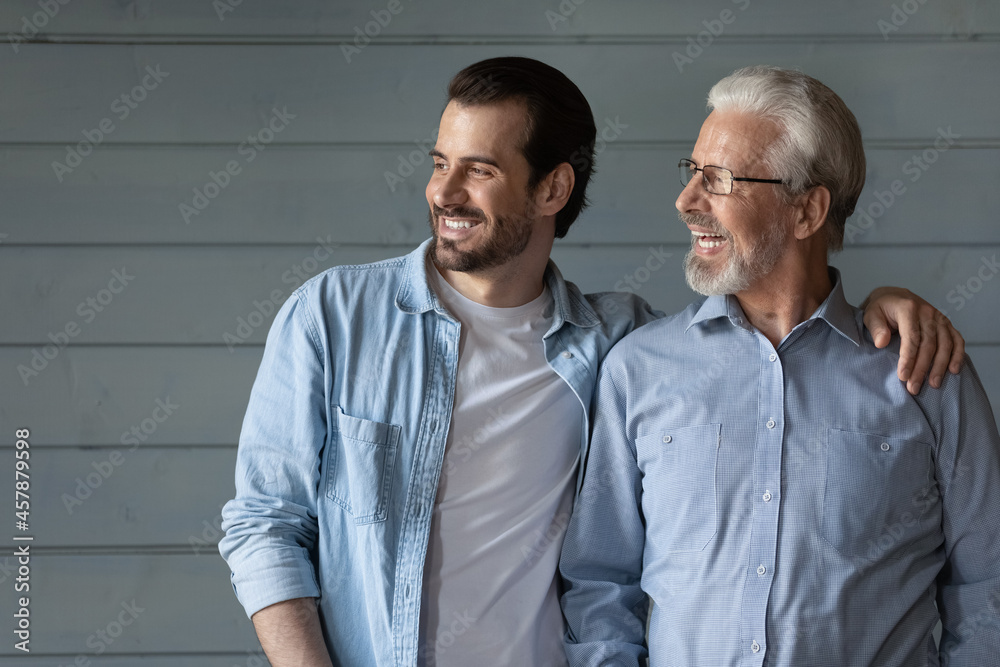 Happy two generations of men hugging, looking in distance, hugging, standing on grey wooden wall background, smiling senior father in glasses and grownup son dreaming, family unity and bonding Happy two generations of men hugging, looking in distance, hugging, standing on grey wooden wall background, smiling senior father in glasses and grownup son dreaming, family unity and bonding
