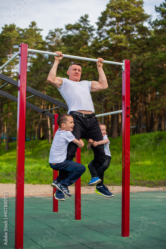 Caucasian man and two boys doing exercises outdoors. The father pulls himself up on the horizontal bar with his sons on the playground.
