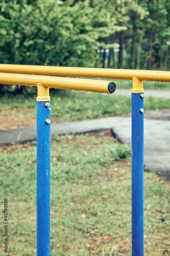 Fototapeta premium Yellow metal bars for exercises with hanging rain drops at sports ground in spring green park on cloudy day extreme close view