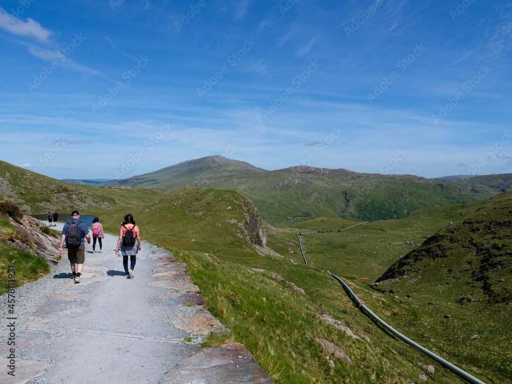 Group of three hikers with backpacks walking on Miners Track back from mountain Snowdon towards Llyn Teyrn lake, water supply pipe for Cwm Dyli hydro-electric power station on the right.