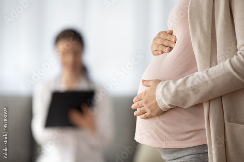 Preparing for baby birth. Close up of woman expecting baby having appointment with doctor at antenatal clinic prenatal healthcare center. Focus on young female holding hands on baby bump in doc office