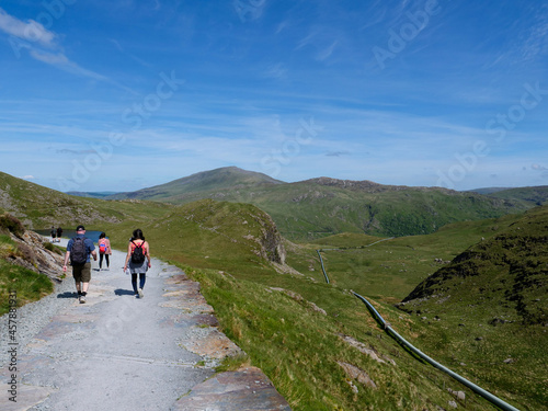 Group of three hikers with backpacks walking on Miners Track back from mountain Snowdon towards Llyn Teyrn lake, water supply pipe for Cwm Dyli hydro-electric power station on the right.
