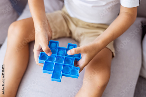 Boy lying on the couch and playing with sensory pop it fidget toy at home. Kid playes stress and anxiety relief fidgeting game. Boy with pop it a heart shape sensory toy.