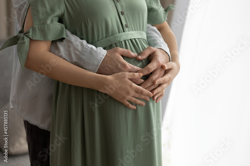 In anticipation. Close up of young male going to be father hug pregnant female from behind hold hands on her big belly. Loving husband embrace beloved wife caring for future mom and baby. Copy space