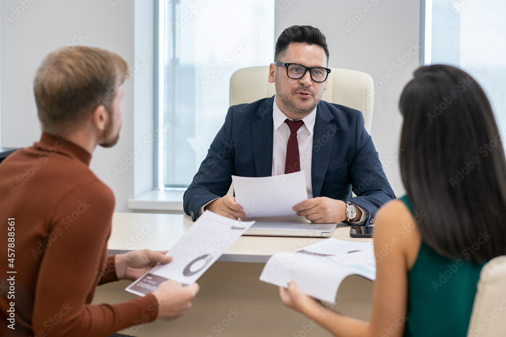 Confident mature businessman with paper discussing financial points or data with his two subordinates Confident mature businessman with paper discussing financial points or data with his two subordinates