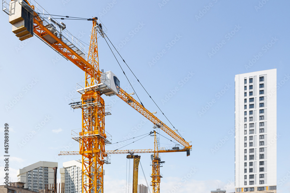 Two yellow tower construction cranes stand against the backdrop of monolithic houses and the blue sky. Construction site and construction of condominiums.