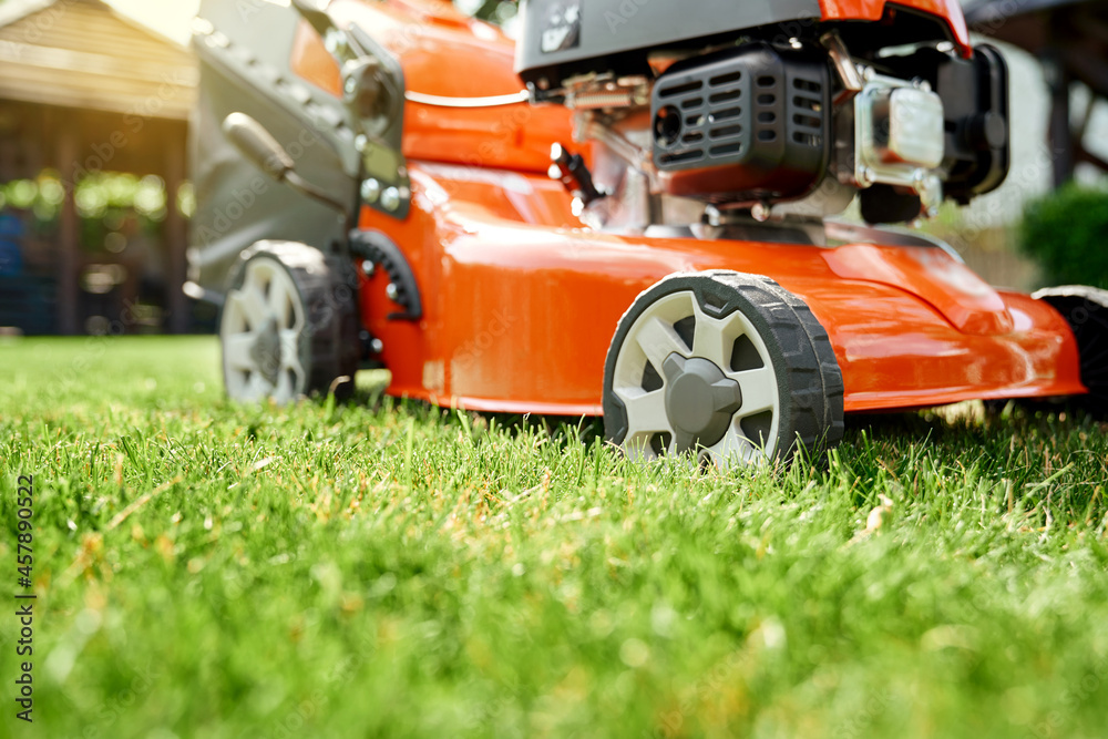 Red lawn mower standing on green grass at summer garden. Close up of modern tool for gardening. Mower grass equipment. Red lawn mower standing on green grass at summer garden. Close up of modern tool for gardening. Mower grass equipment.
