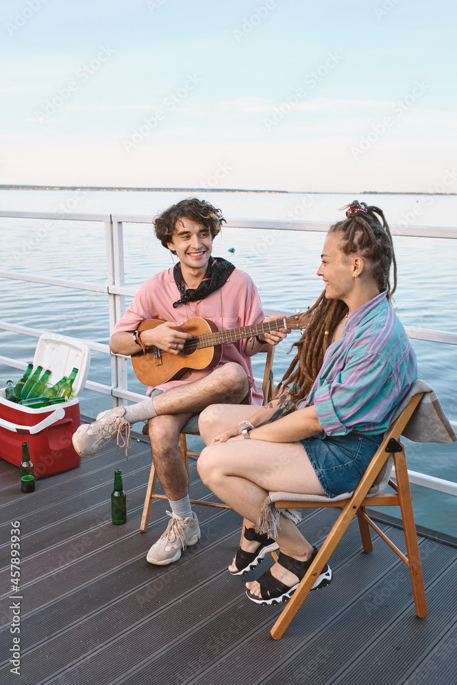 Happy young man with guitar looking at his girlfriend while both relaxing on pier on summer day