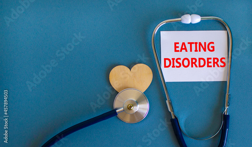 Eating disorders symbol. White card with words Eating disorders, beautiful blue background, wooden heart and stethoscope. Medical and eating disorders concept.
