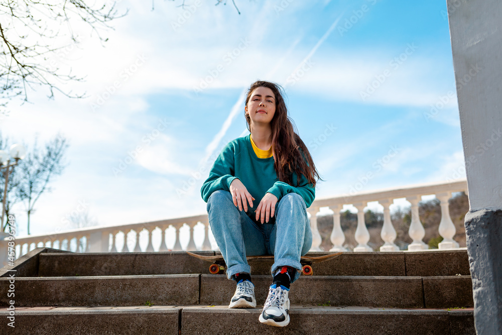 Portrait of a cute Caucasian teen girl sitting on a skateboard. In the background, the blue sky and the Boulevard. Bottom view. Copy space. Concept of sports lifestyle and street culture