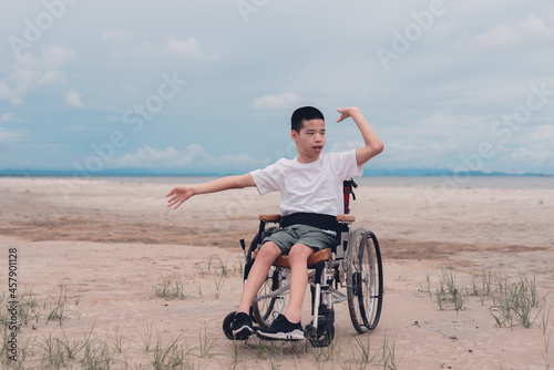 Happy handicapped teenager boy in wheelchair holding red cap and smiling face,Sea beach background, Lifestyle happy disabled child travel , Good mental health concept