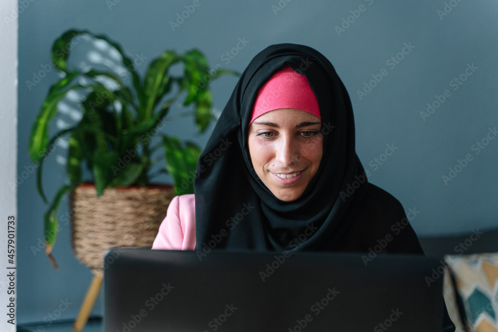 Young muslim woman wearing a hijab using technology indoors. Horizontal view of arabic woman working with laptop at home. Technology and muslim women lifestyle.