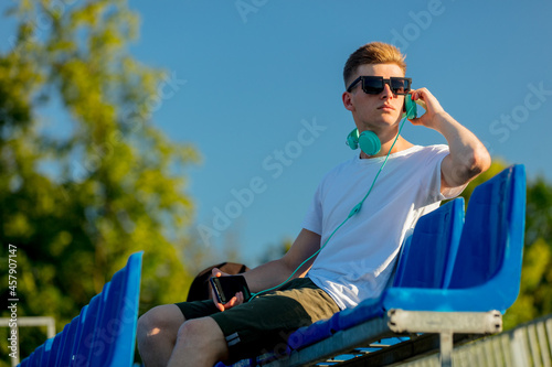 Young teen boy in sunglasses with headphones sitting in blue sit on stadium tribune