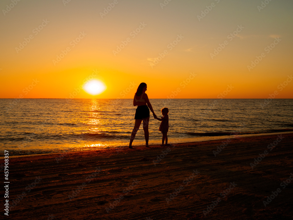A young woman and a little baby girl hold hands on the seashore against the backdrop of the setting sun.