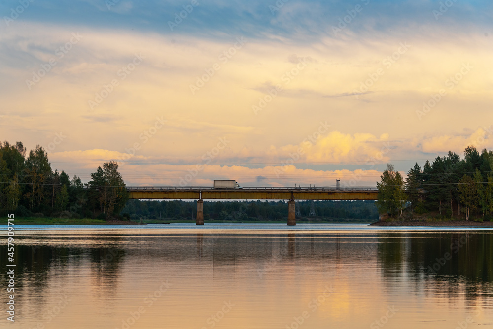 Landscape with a river and a forest illuminated by the setting sun. Reflection of clouds in the water. Suburban road bridge through the river.