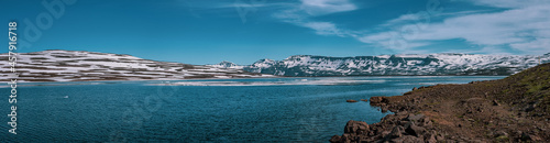 Panoramic frog view of Heidharvatn lake in eastern iceland, glacial lake with some blocks of ice floating and majestic mountains and road next to it.