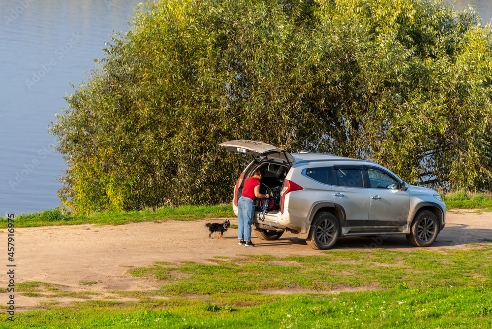 Mature woman with a dog loads the trunk of her car on the river bank.