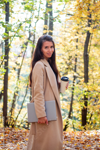 Brunette woman in autumn park with laptop and cup of coffee. Student. Teacher. Online education.