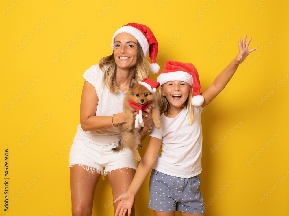 Obraz premium Happy mother and daughter with small dog in santa hat over yellow background. Christmas season concept.