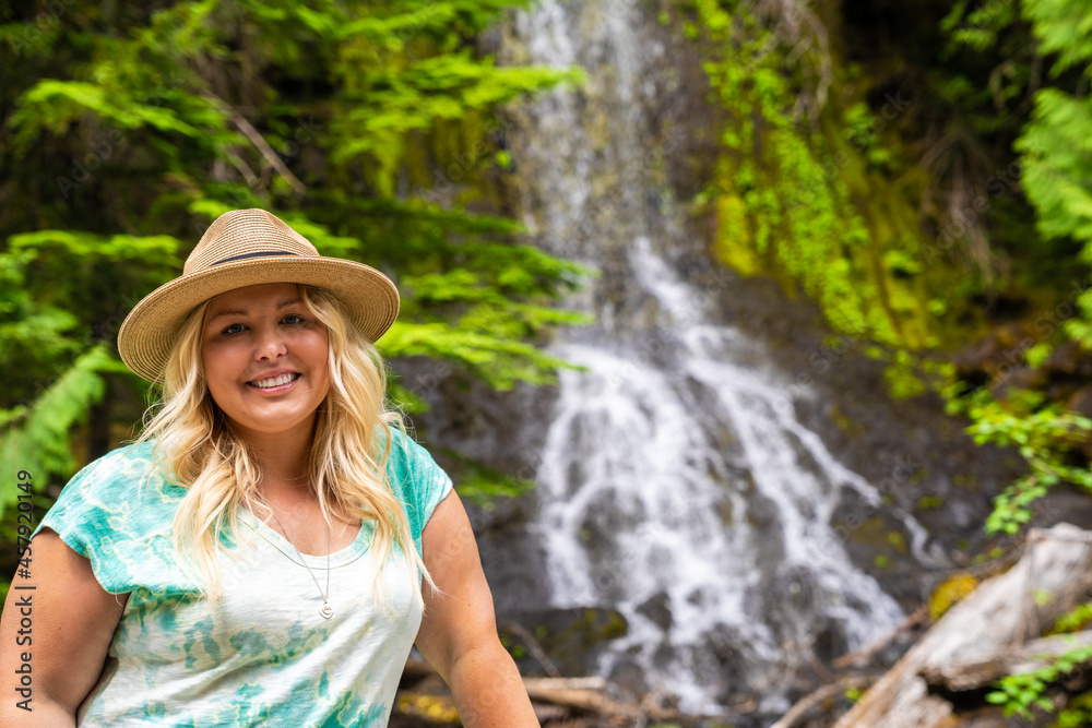 Cute blonde woman wearing a hat poses in front of Falls Creek waterfall in Mt. Rainier National Park