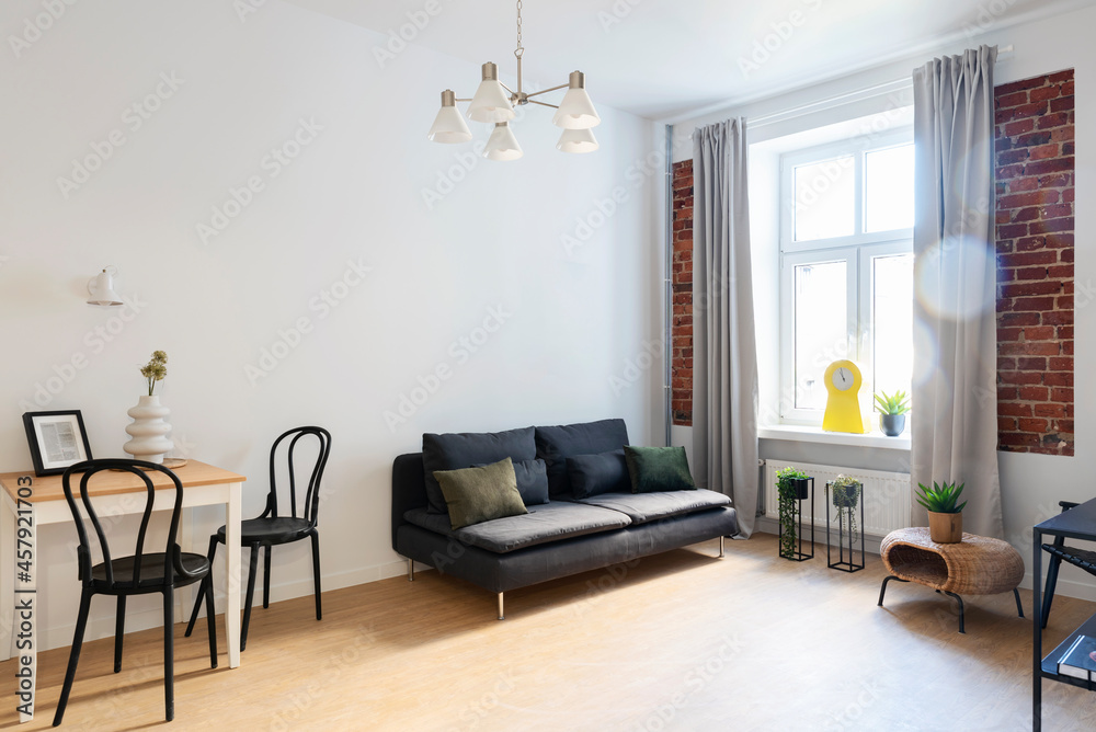Fototapeta premium Interior of living room in loft apartment in industrial design with window and brick wall. Comfortable sofa with pillows and table with stylish chair. Hotel.