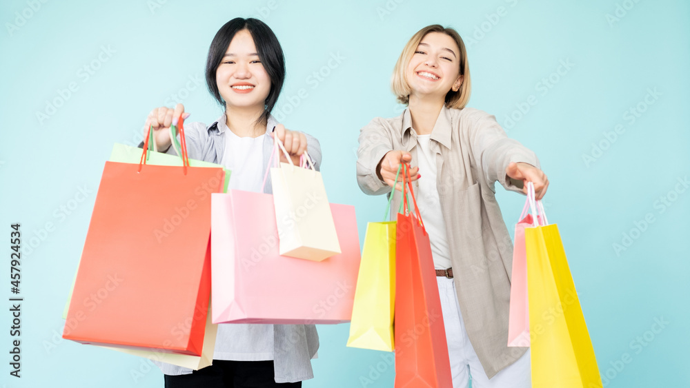 Shopaholic lifestyle. Happy women. Hot sale. Best offer. Shopping discount. Pretty smiling asian caucasian friends showing colorful purchase bags isolated blue. Shopaholic lifestyle. Happy women. Hot sale. Best offer. Shopping discount. Pretty smiling asian caucasian friends showing colorful purchase bags isolated blue.