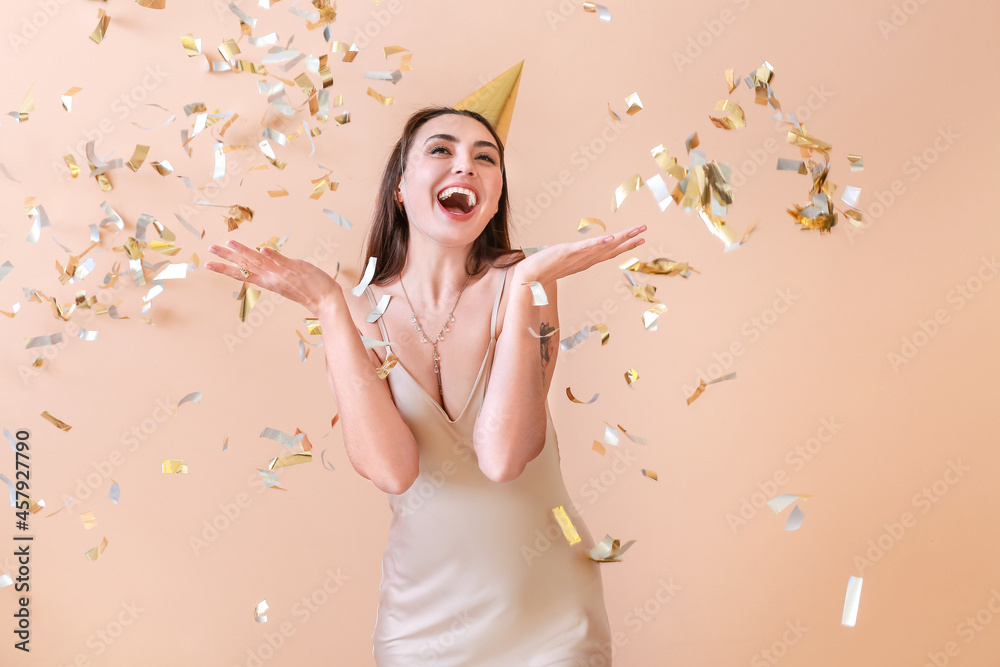 Fototapeta premium Happy woman in party hat and with flying confetti on color background