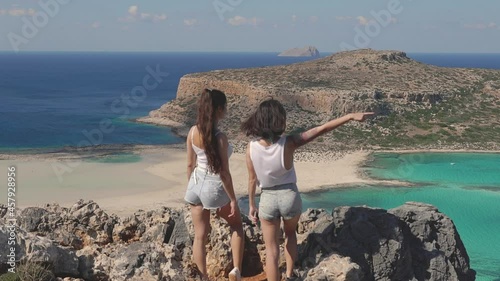 Two beautiful girls standing on edge of cliff. Female friends admire view of blue sea laggon and rocky beach, pointing in different directions and enjoy summer. Concept of travel, vacation, adventure photo