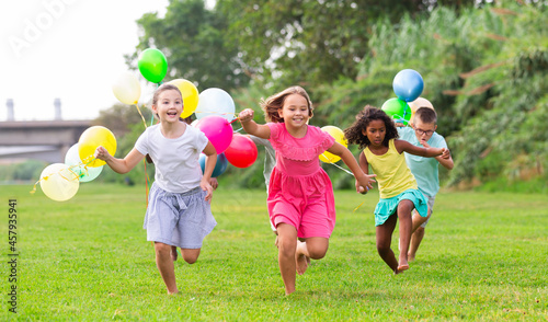 Barefoot children running through field with balloons in hands and smiling.
