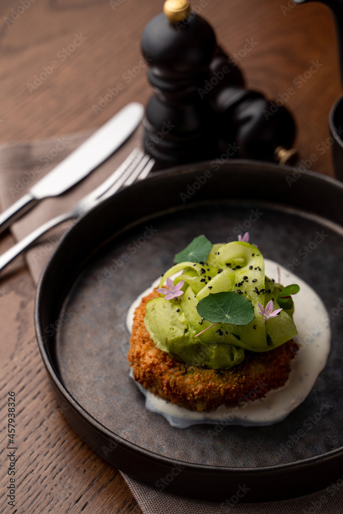 Menu photography in a dark restaurant: roasted fish cake with avocado and garlic sauce on a black dish, soft light