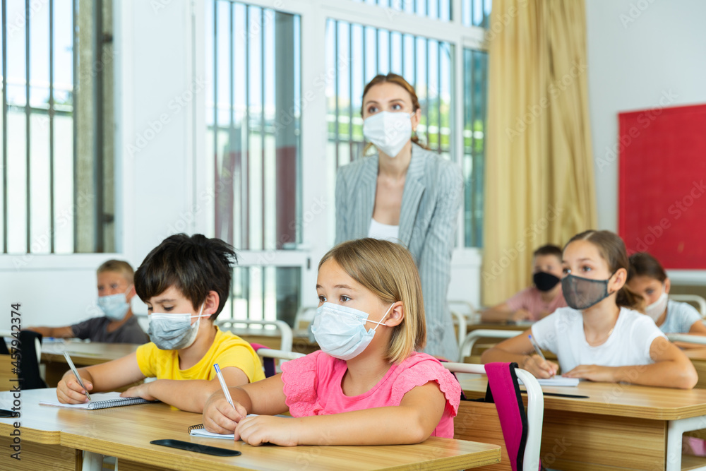 Fototapeta premium School boy and girl in face masks listening to teacher and writing exercises in notebook at lesson in primary school
