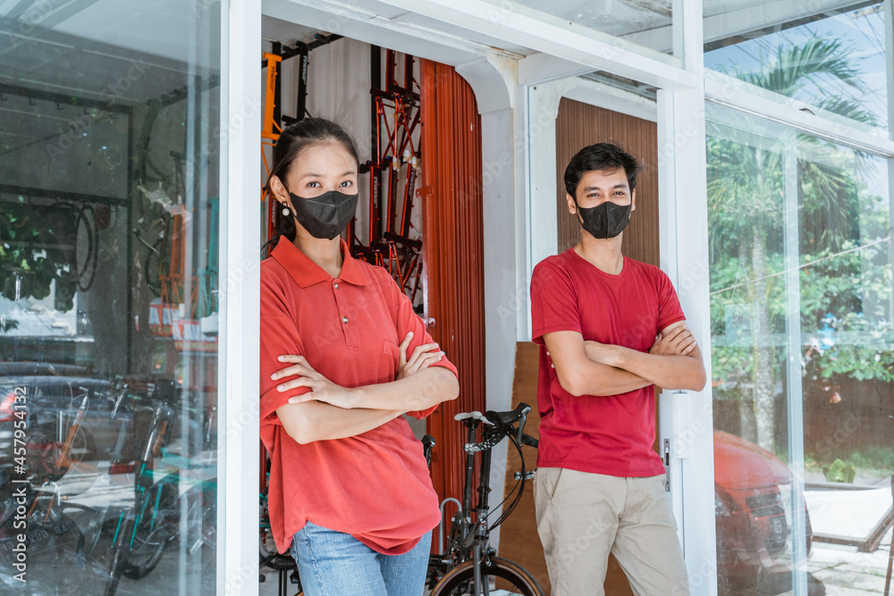 Salesman and saleswoman in mask stands leaning in front of the glass door with folded hands