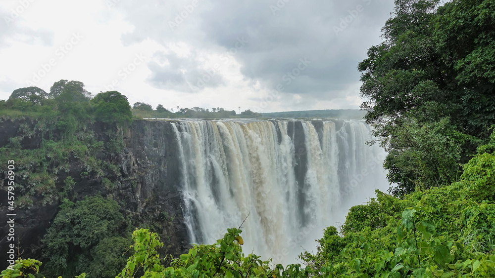 The Zambezi River flows from the edge of the plateau into the gorge in powerful streams and forms Victoria Falls. There is a fog over the abyss. In the foreground, lush green vegetation. Zimbabwe