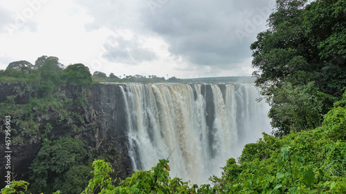 The Zambezi River flows from the edge of the plateau into the gorge in powerful streams and forms Victoria Falls. There is a fog over the abyss. In the foreground  lush green vegetation. Zimbabwe