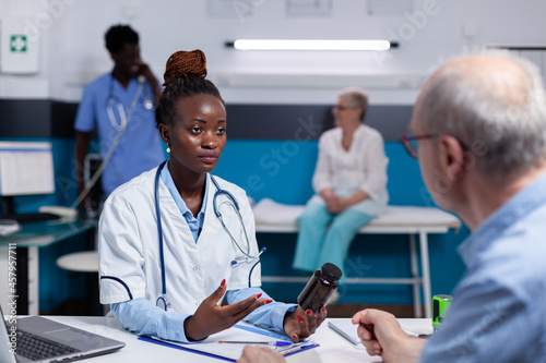 Black woman with doctor profession holding bottle of pills in medical cabinet explaining treatment to senior patient. African american man nurse talking to old person in background